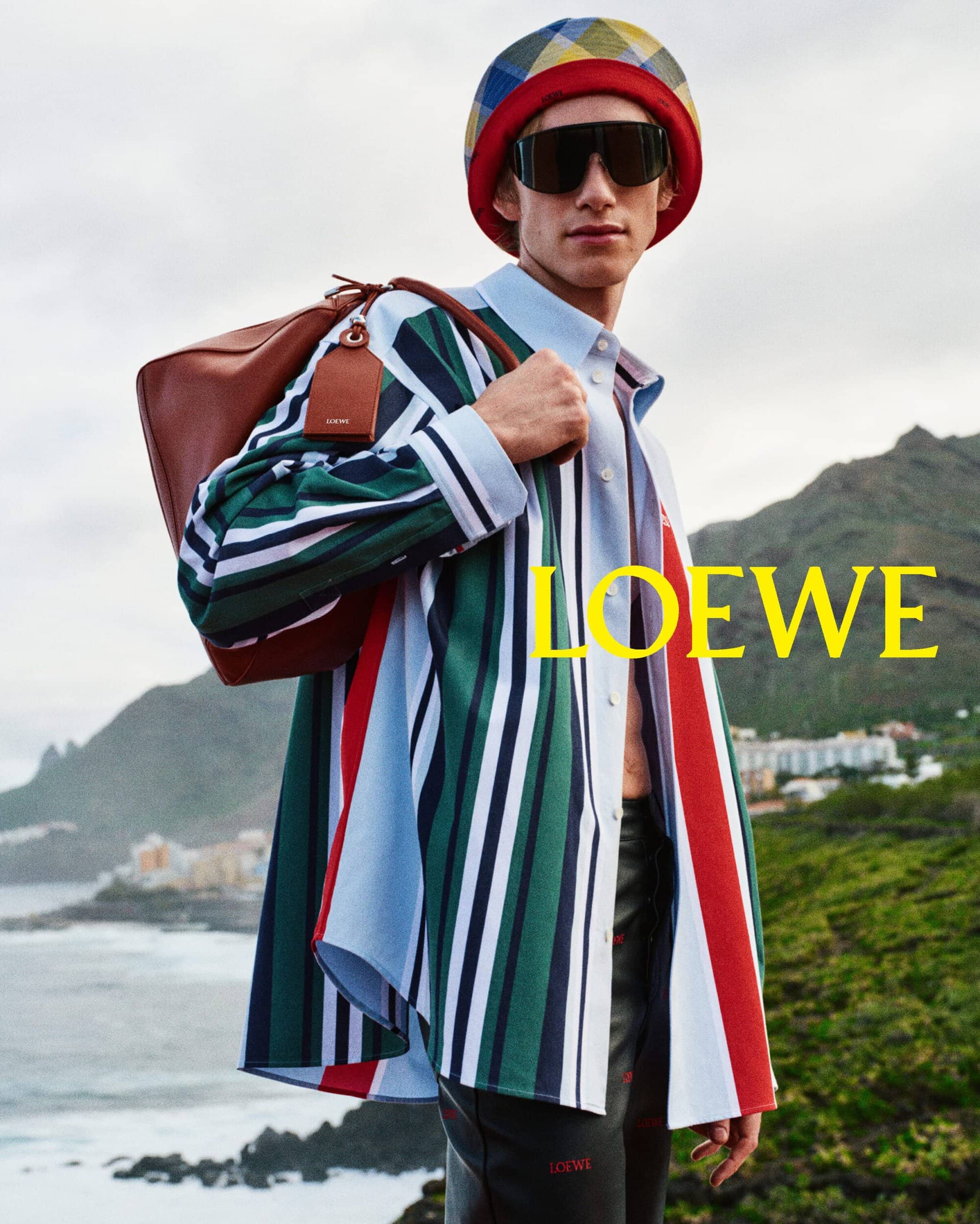 Model in colourful striped shirt and patterned bucket hat carrying a brown leather shoulder bag against a coastal mountain backdrop
