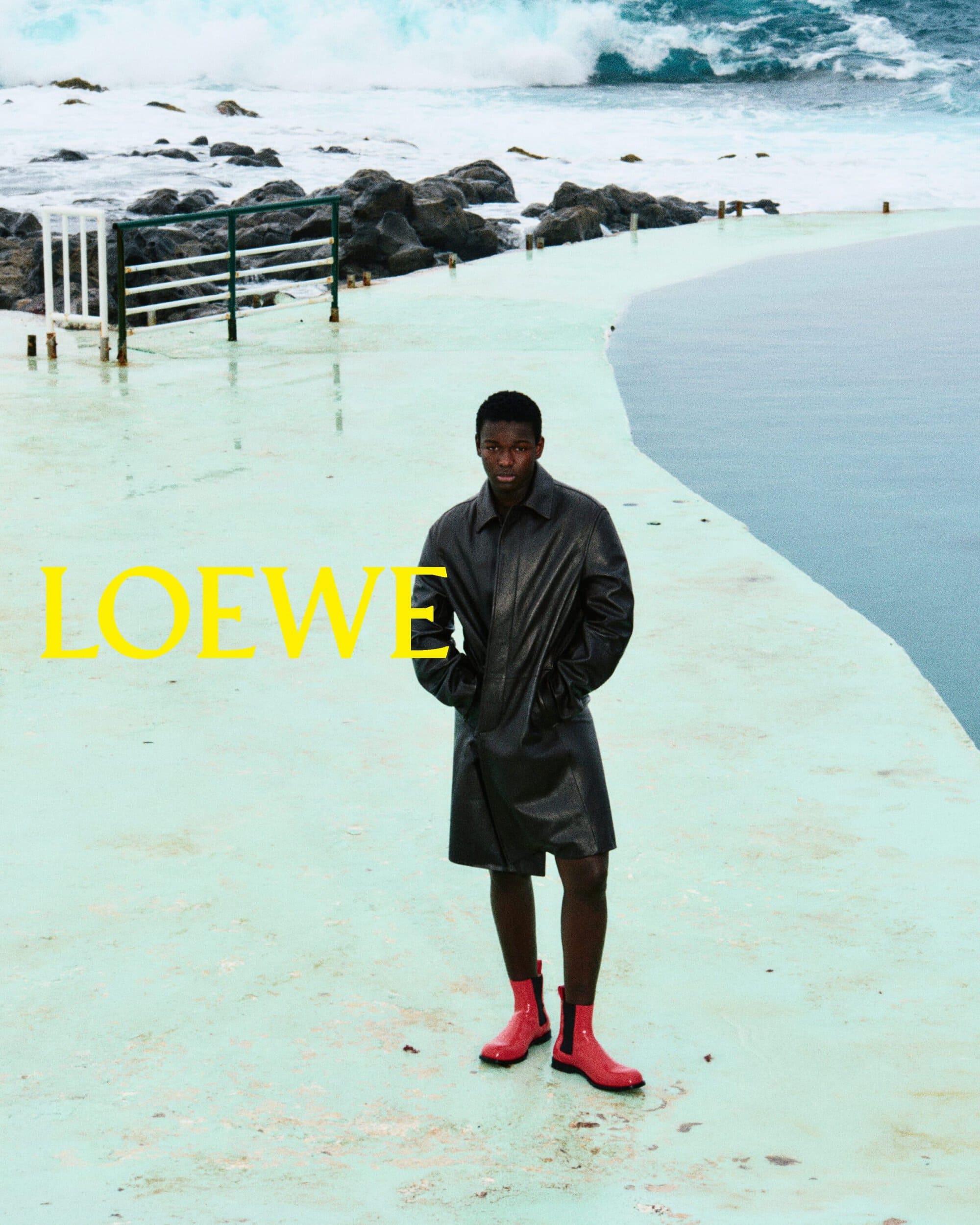 Man wearing a dark coat and red boots standing on a pale coastal promenade with ocean waves in background