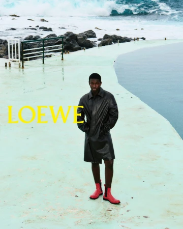 Man wearing a dark coat and red boots standing on a pale coastal promenade with ocean waves in background