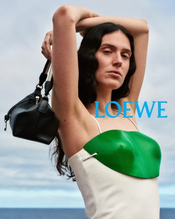 Model with arms raised holding a black leather handbag against an overcast sky and water background