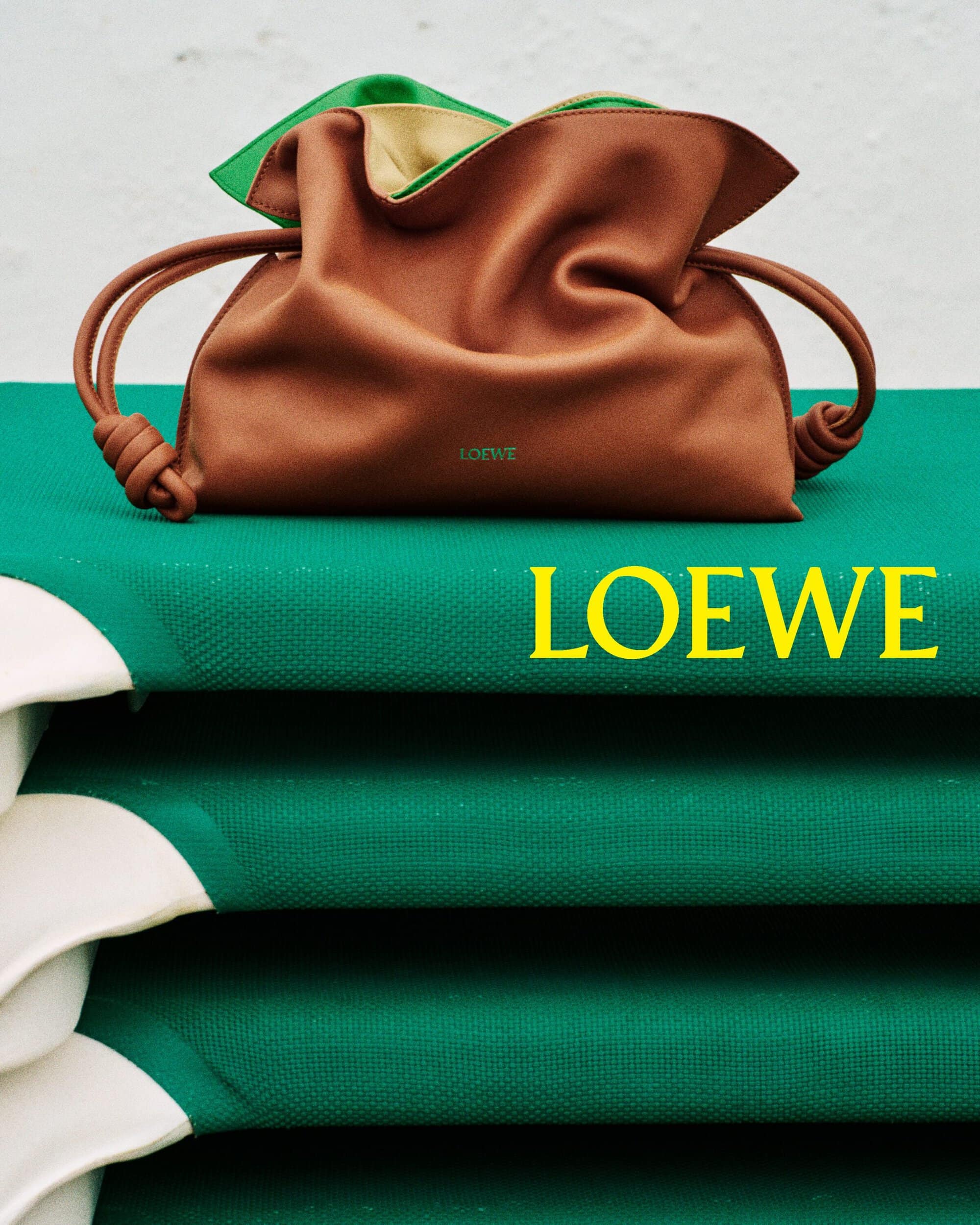 Brown leather Loewe handbag resting on a stacked green and white textured surface with bold yellow typography.