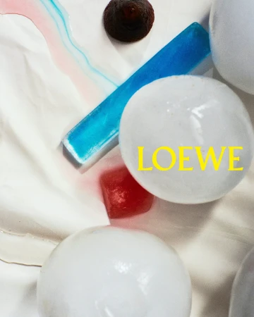 Close-up abstract still life with white spheres, blue rectangular resin block, red cube, and yellow LOEWE branding.