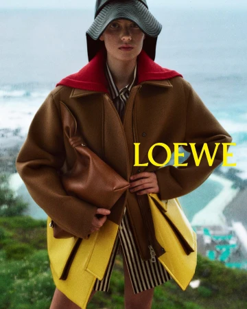 Model wearing a brown coat and hat holding a brown leather bag against an ocean background