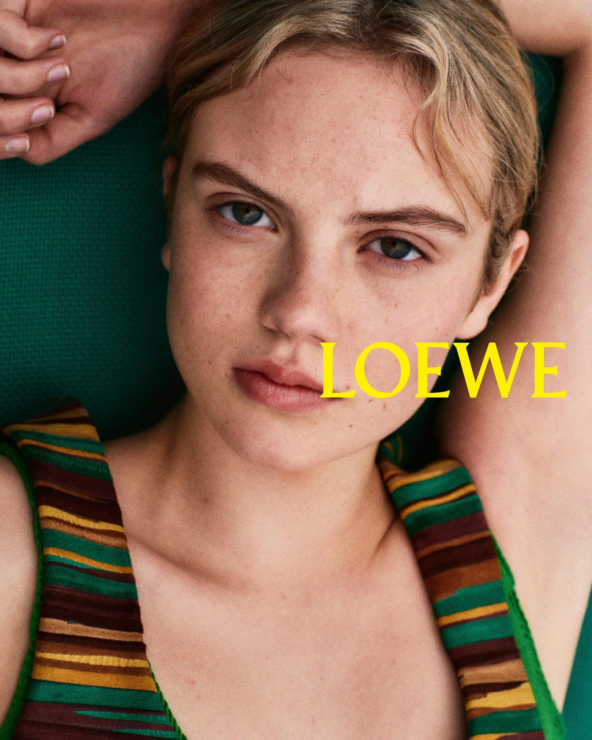 Close-up portrait of a young woman wearing a striped green and brown top with her hands behind her head against a green background