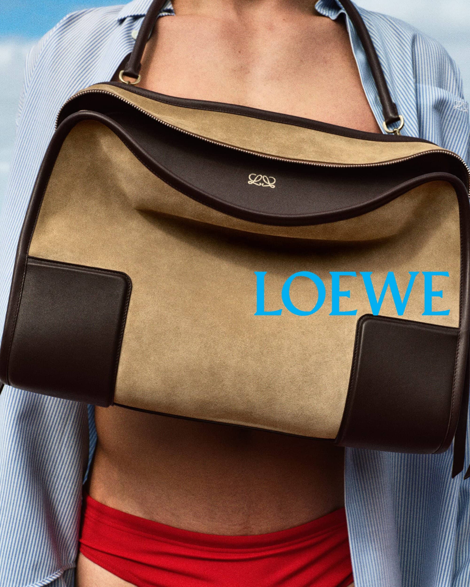 Close-up of a model holding a large tan and brown suede Loewe handbag against a clear blue sky.