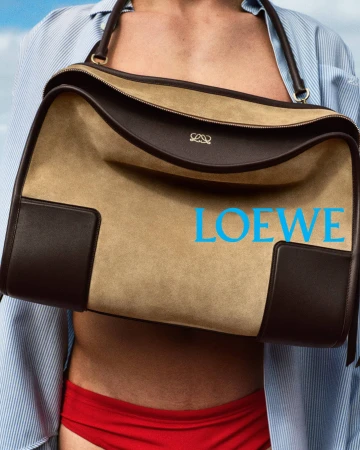 Close-up of a model holding a large tan and brown suede Loewe handbag against a clear blue sky.