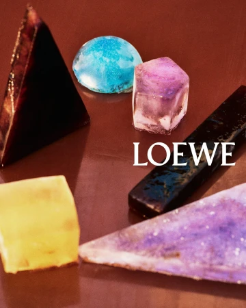 Close-up of geometric resin and stone objects in blue, yellow, and purple on a brown surface with Loewe text