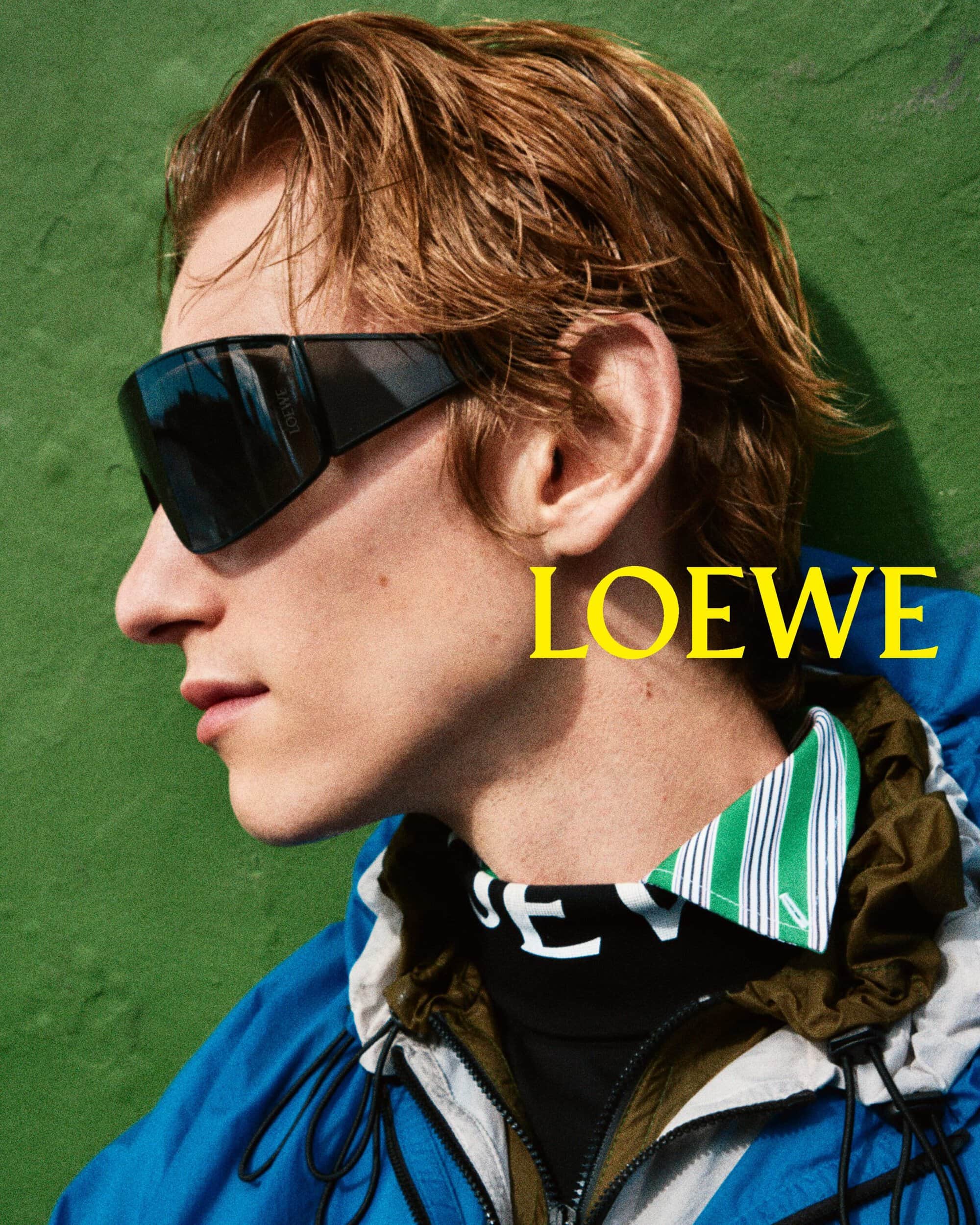 Profile portrait of a young man wearing black sunglasses, a blue jacket, and a striped collar against a textured green background.