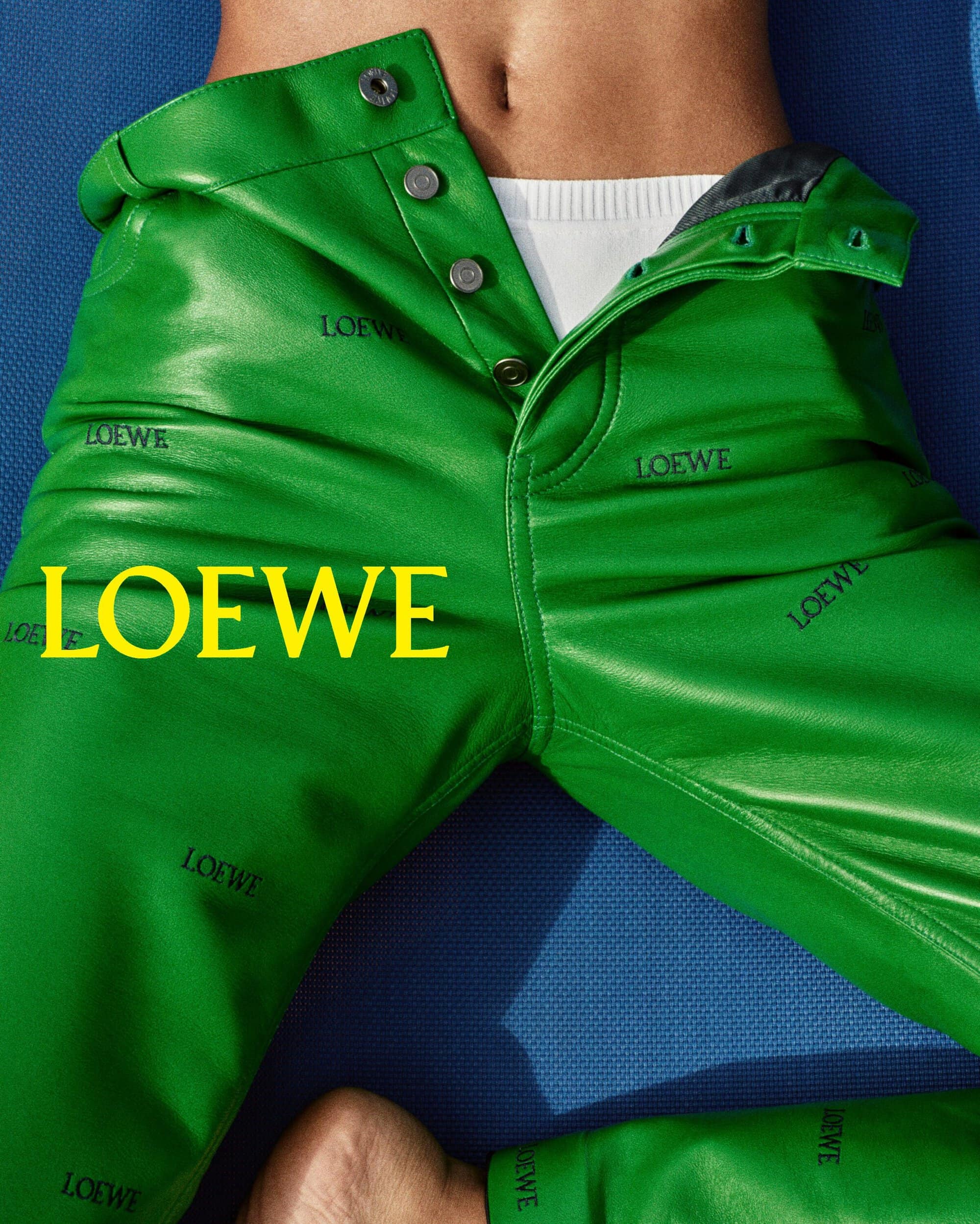 Close-up of vibrant green leather trousers with button fly and repeating Loewe logo print against blue textured background