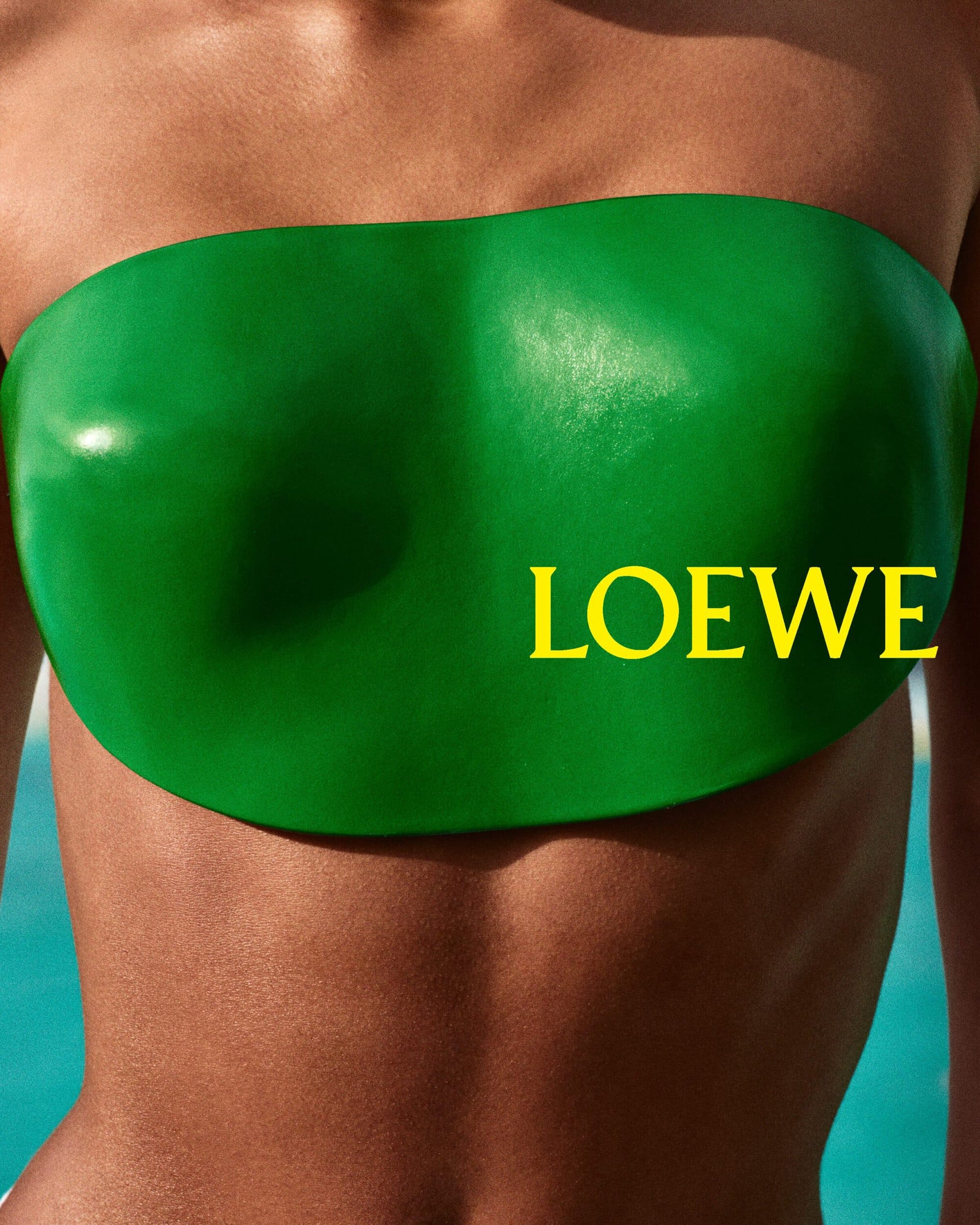 Close-up studio shot of an adult female model wearing a green strapless top with the Loewe logo in yellow text over a blue background.
