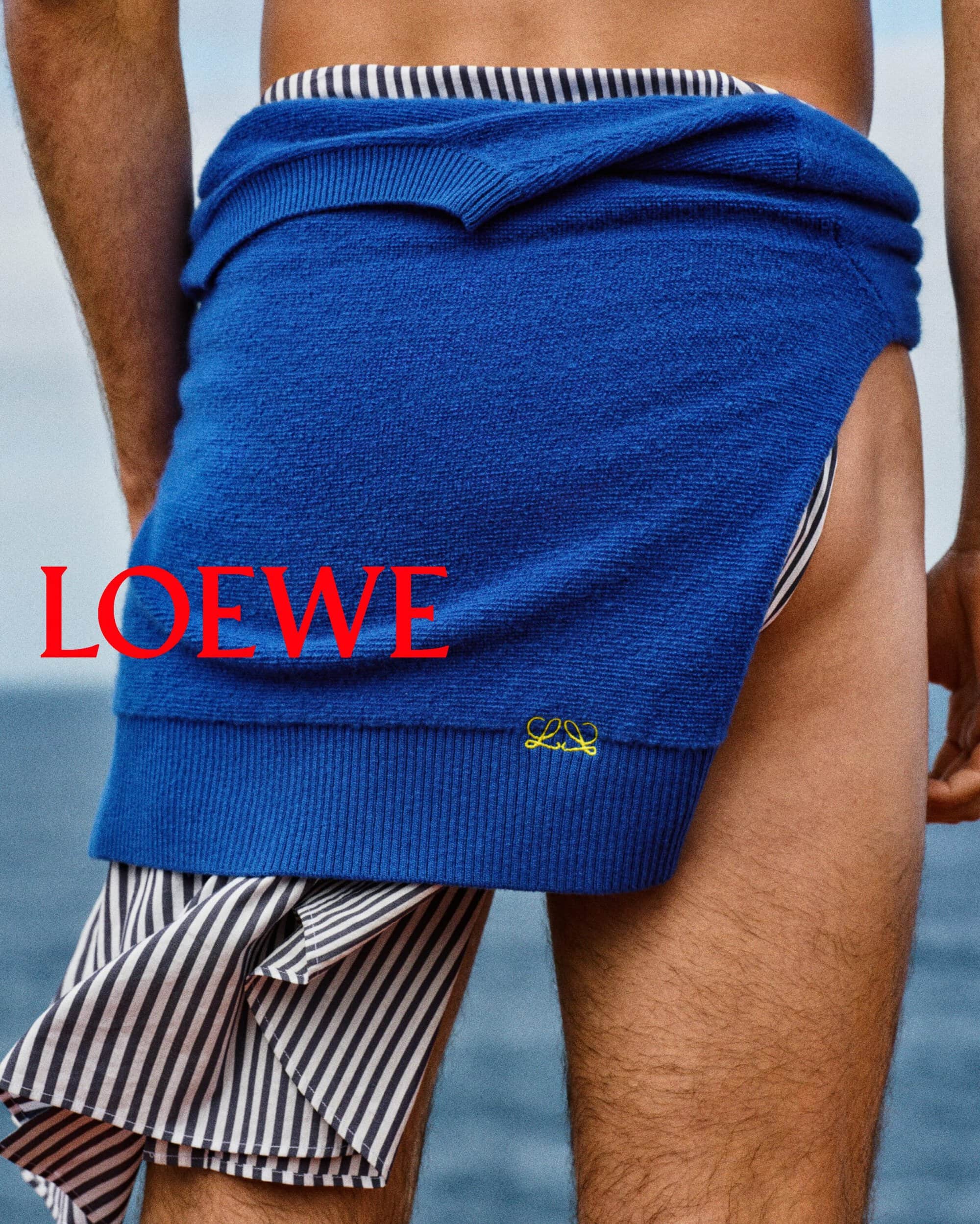 Close-up rear view of a person wearing a royal blue sweater tied around the waist over striped shorts against an ocean background.