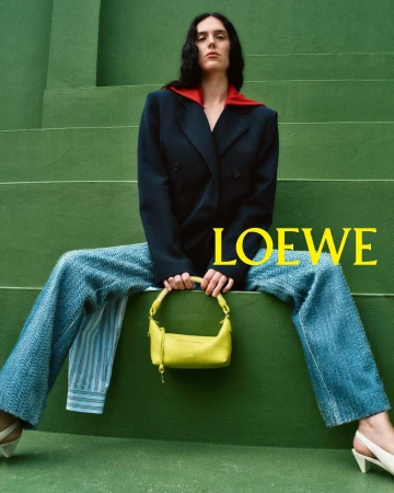 Model seated on green steps wearing dark blazer and denim trousers, holding a bright yellow handbag.