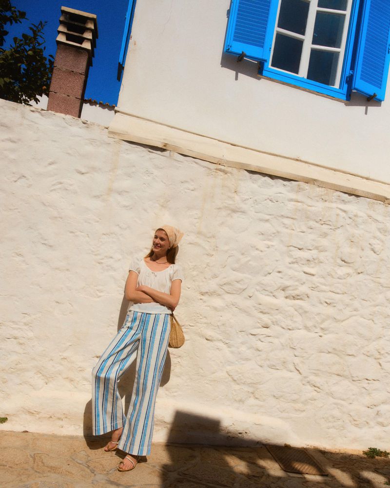 Model leaning against a textured stone wall wearing striped trousers and a white top under a bright blue window shutter.