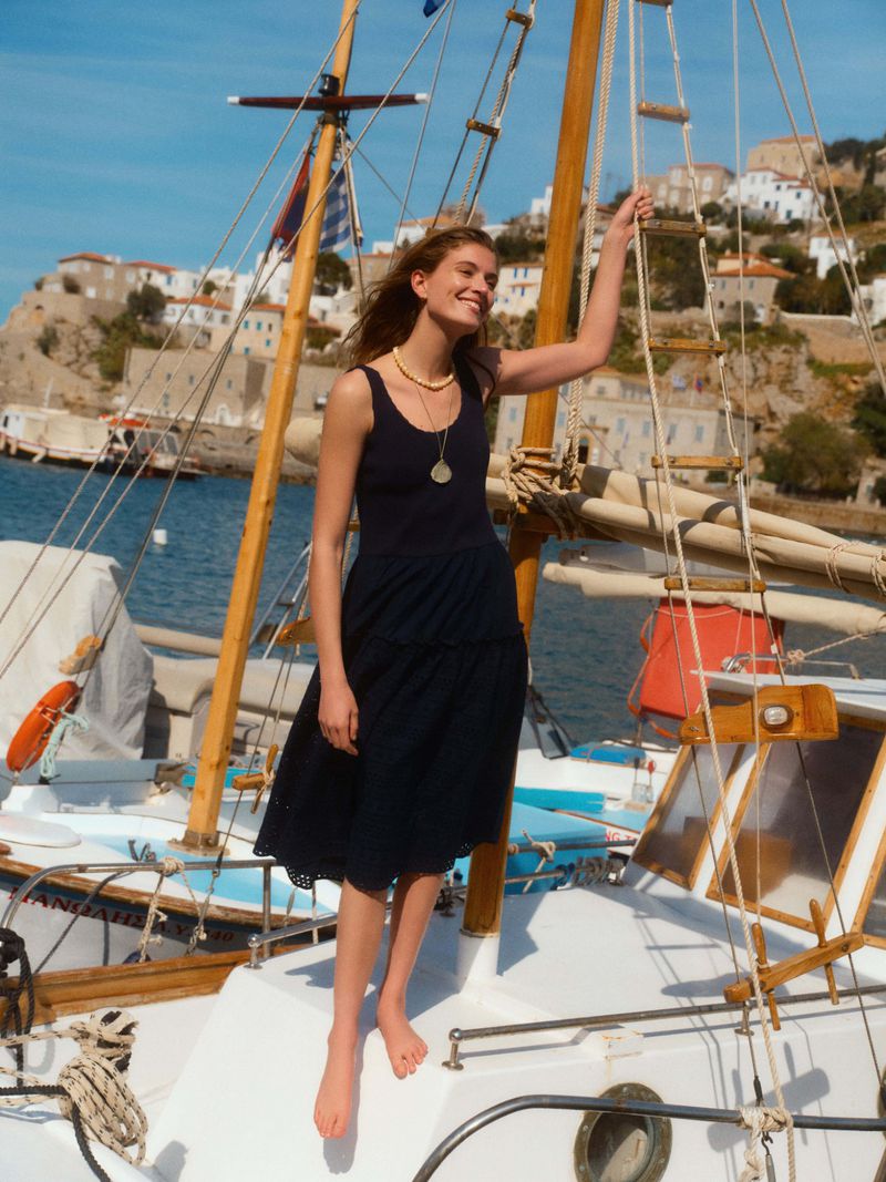 Model standing barefoot on a sailboat deck wearing a dark sleeveless dress against a coastal village background.
