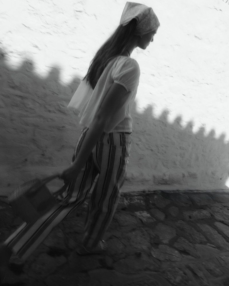 Model walking outdoors wearing a headscarf, white blouse, and striped trousers carrying a basket bag