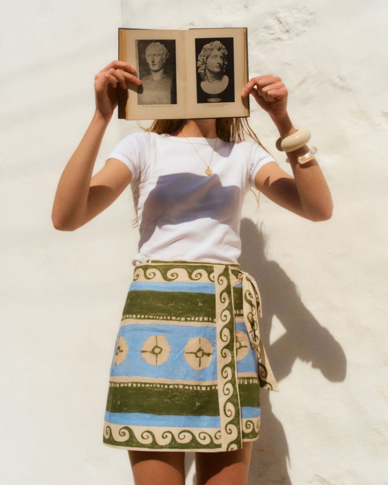 Model holding an open vintage book over her face, wearing a white t-shirt and patterned blue and green skirt against a white wall.