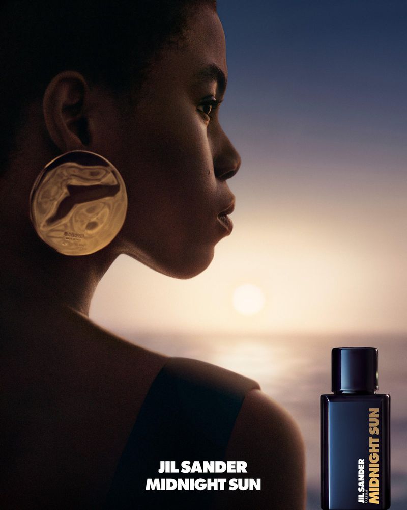 Profile of woman with large gold earring against a sunset. A bottle of JIL SANDER MIDNIGHT SUN perfume is in the foreground.
