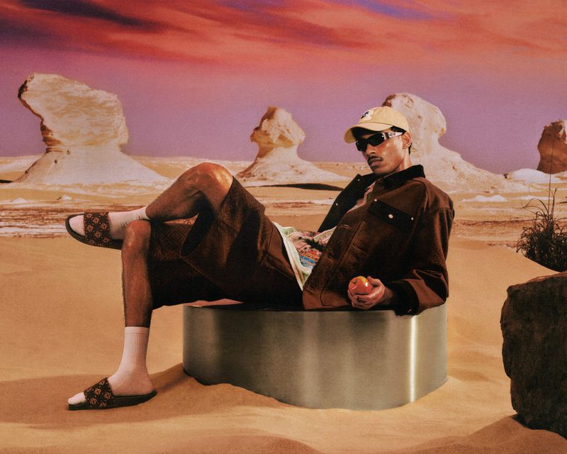 Model seated on a metal cylinder in a desert landscape wearing a brown jacket, shorts, cap, and sunglasses.