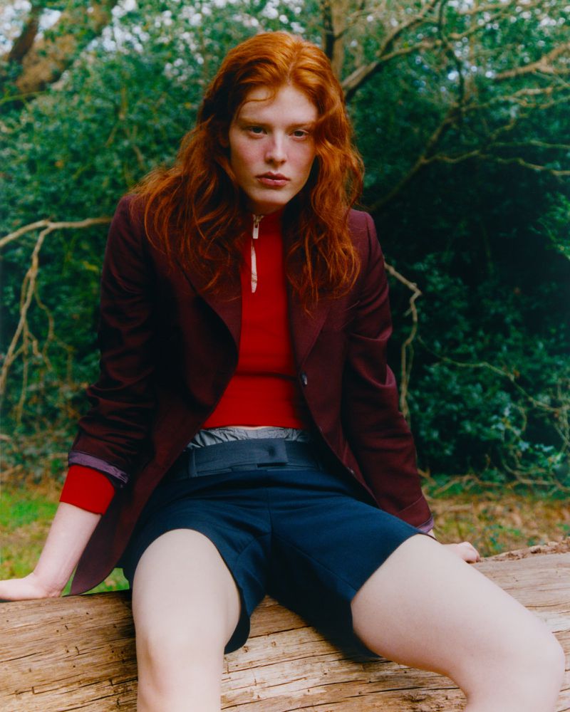 Young redhead poses on a log in a burgundy jacket and navy shorts, set against a backdrop of lush green foliage, outdoors.