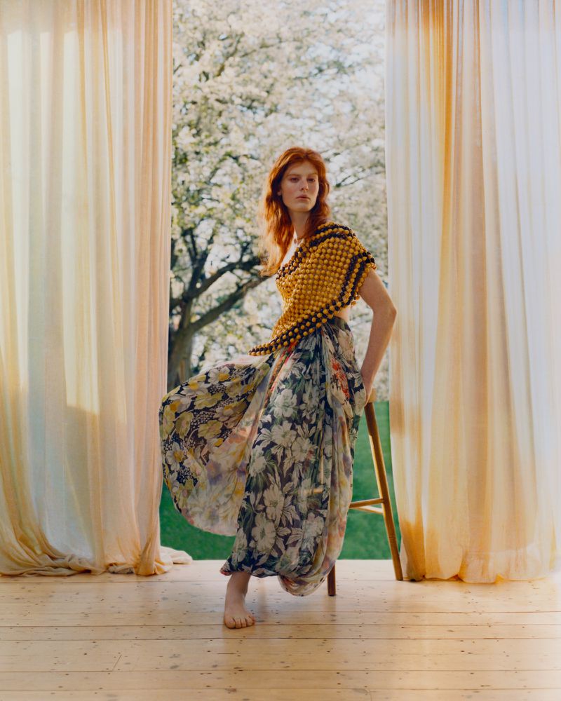 Red-haired woman in a floral skirt and yellow top sits barefoot on a stool, framed by curtains and a tree in full bloom.