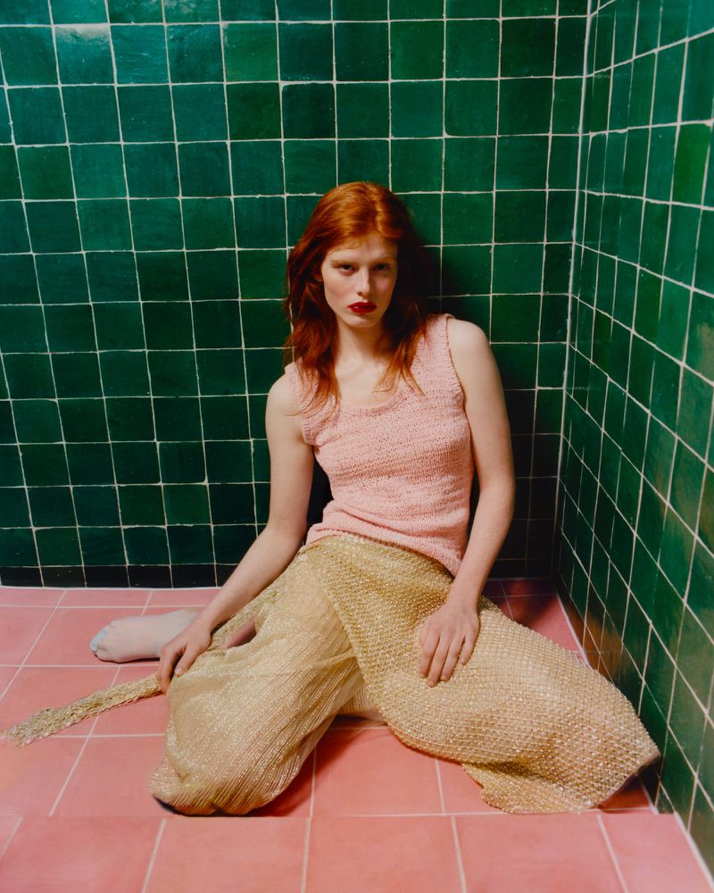 Red-haired woman poses in a pink top and gold, flowing pants against a green and pink tiled backdrop, fashion photography.