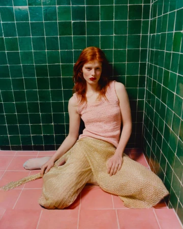 Red-haired woman poses in a pink top and gold, flowing pants against a green and pink tiled backdrop, fashion photography.