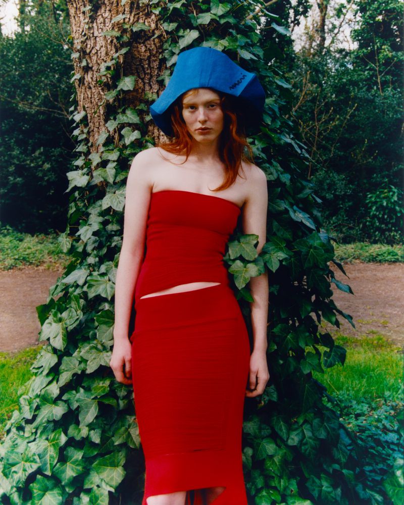 Adult woman in red strapless dress and blue hat poses by a tree in an outdoor green environment with natural lighting.