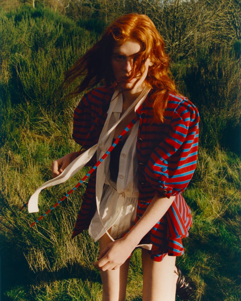 Portrait of a red-haired woman in a striped top outdoors. She is standing in a grassy field with trees in the background.