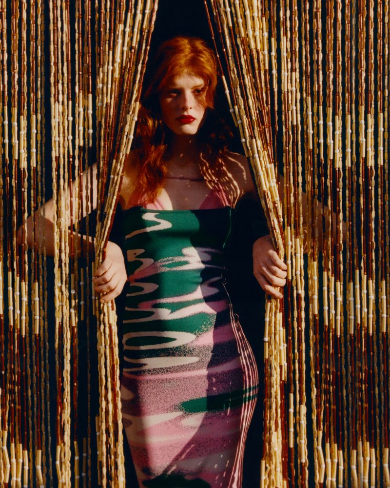 Redhead woman in patterned dress poses between beaded curtains in a fashion portrait with warm lighting and soft focus.