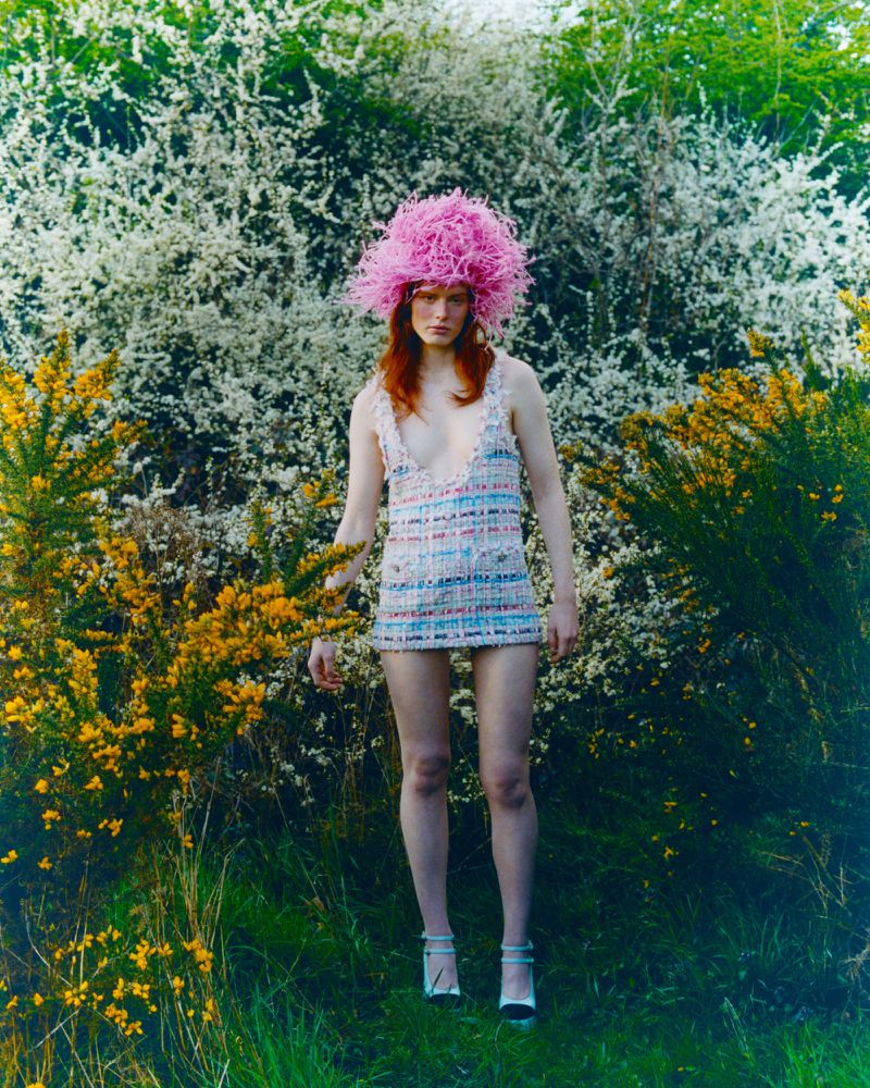 A young woman with red hair wears a textured mini dress and a bright pink hat made of feathers while standing in a grassy field.