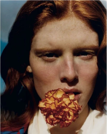 Close-up portrait of a red-haired woman holding a flower in her mouth, against a soft blue and white gradient background.