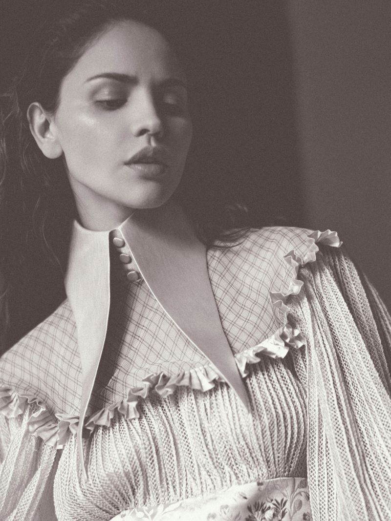 Portrait of an adult woman with dark hair wearing a vintage-style dress with ruffles and a high collar, photographed indoors in muted tones.