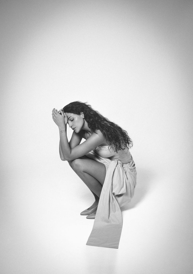 Black and white editorial portrait of Eiza Gonzalez crouching against a seamless white studio background