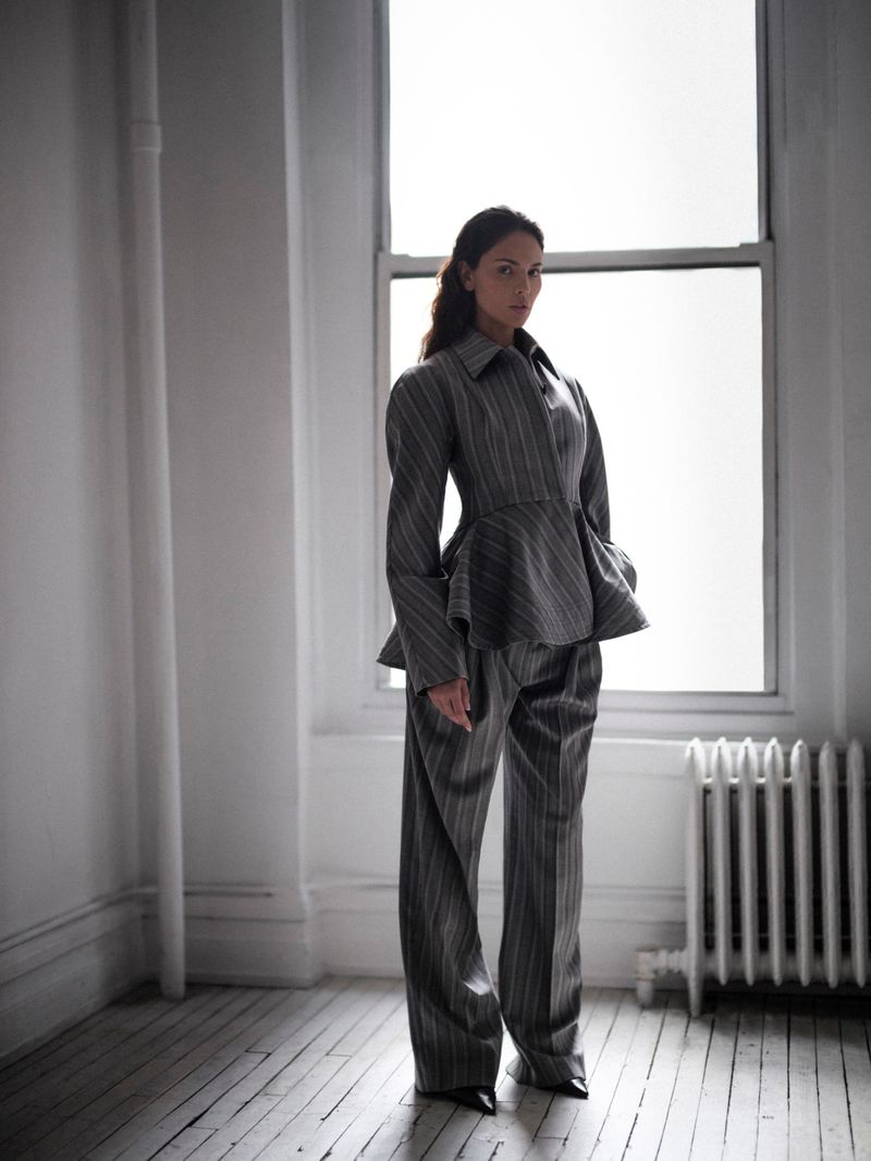 Eiza Gonzalez wearing a striped grey suit standing in a room with a large window and white wooden floor.