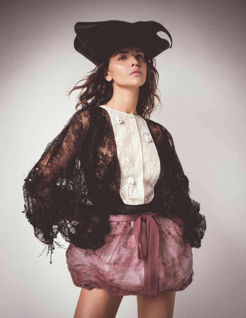 Eiza Gonzalez wearing a black lace jacket, white bib shirt, pink bubble skirt, and wide-brim hat against grey backdrop