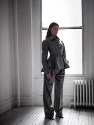 Eiza Gonzalez wearing a striped grey suit standing in a room with a large window and white wooden floor.