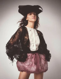 Eiza Gonzalez wearing a black lace jacket, white bib shirt, pink bubble skirt, and wide-brim hat against grey backdrop