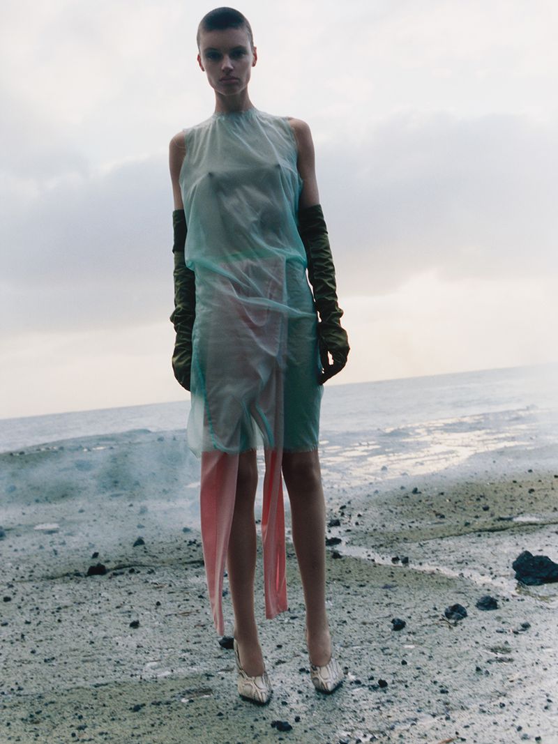 Fashion shot of a woman with a shaved head wearing a translucent dress and gloves, standing on a shore with a cloudy sky and ocean backdrop.