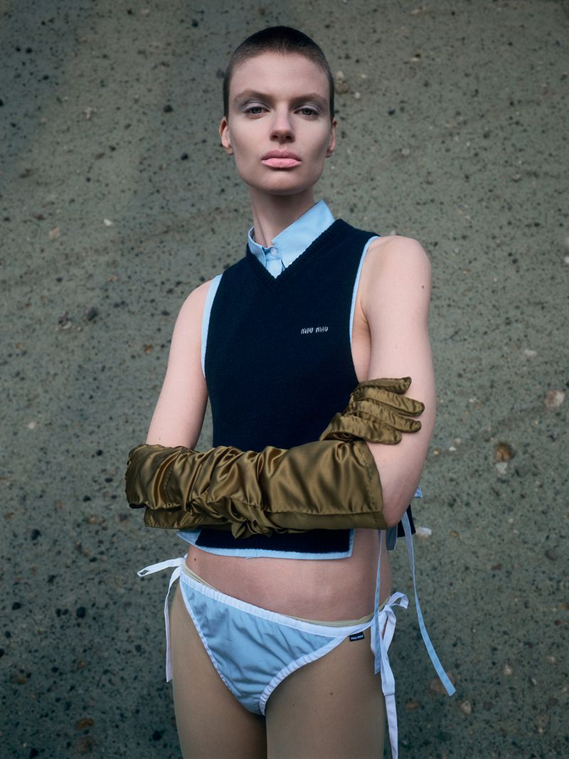 Model with a shaved head wearing a dark sweater vest, light blue briefs, and long gold satin gloves against a concrete wall