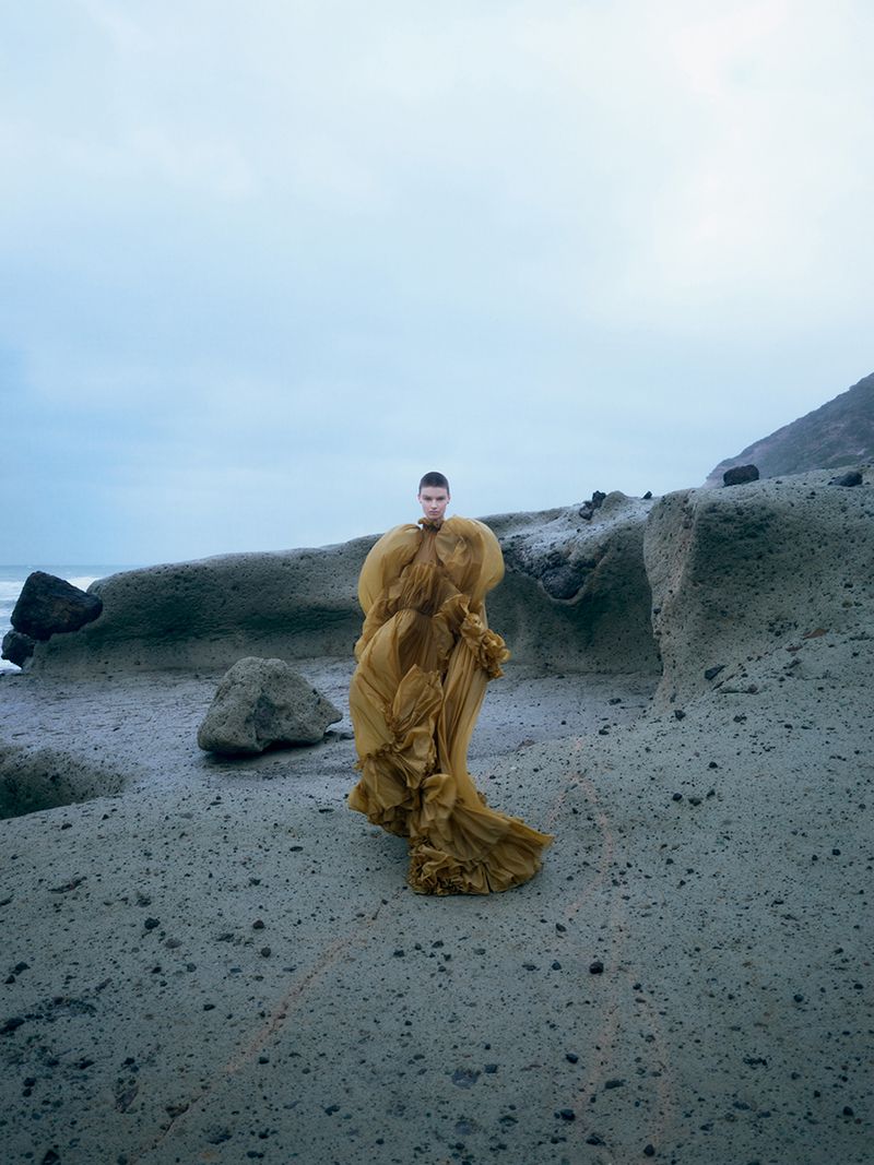 Model standing on a rocky coastal shore wearing an oversized yellow tulle dress under an overcast sky.