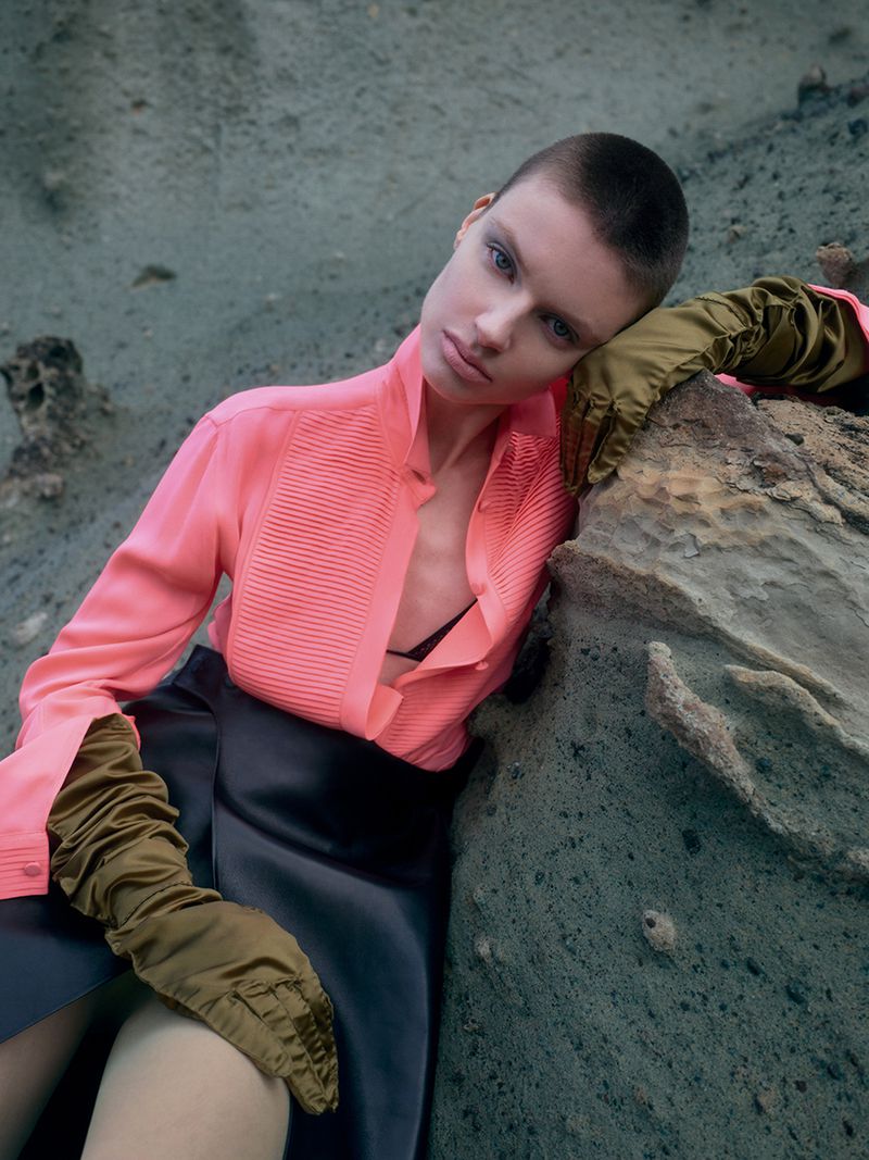 Model with buzzcut wearing a coral pink blouse and olive gloves leaning against a rocky terrain background