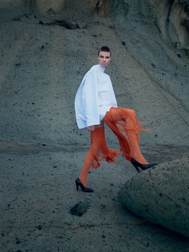 Model with short hair wearing a white top and orange fringe skirt stepping on a rocky slope.