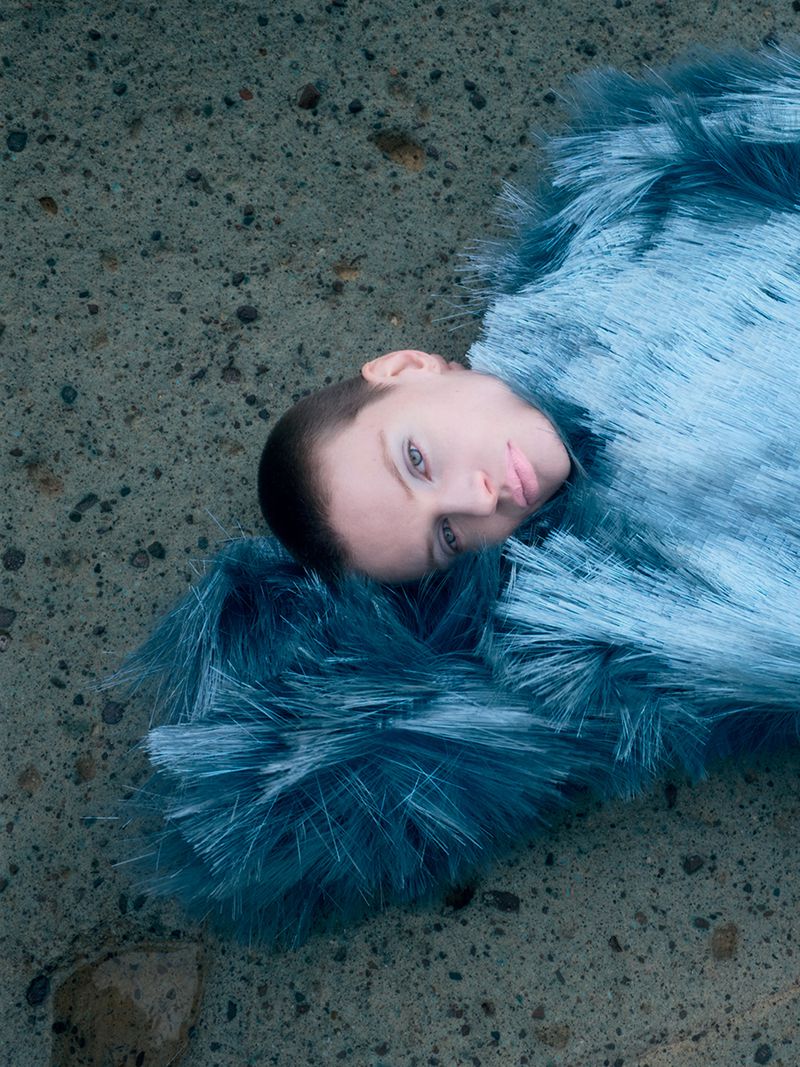 Model with short hair lying on rough stone ground wearing a textured blue fringed garment
