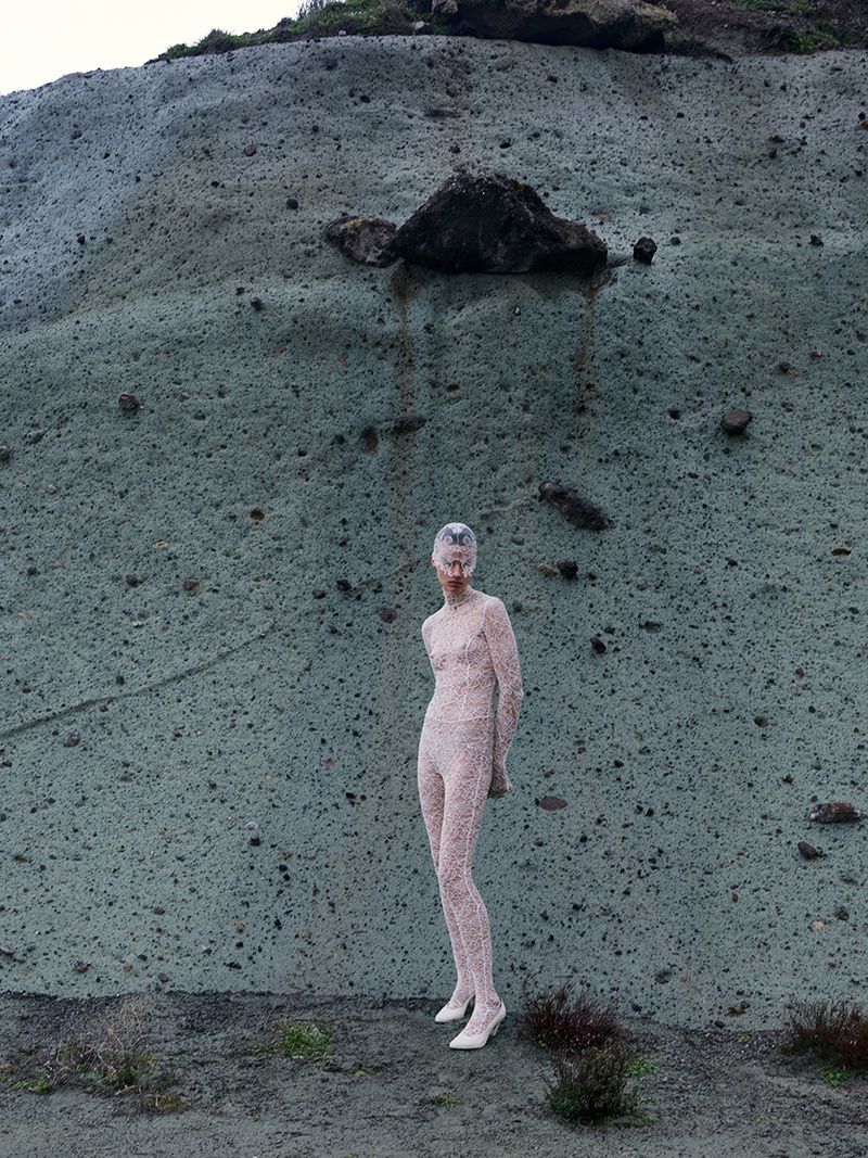 Model wearing patterned lace bodysuit standing in front of a textured stone cliff face