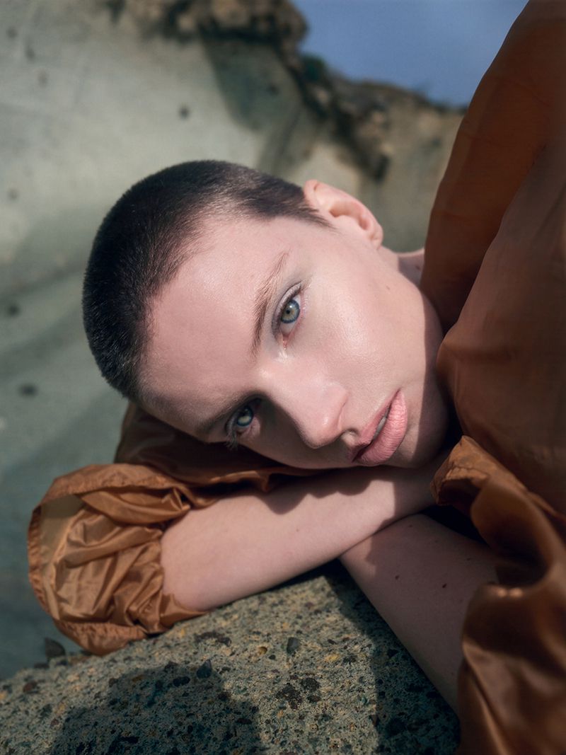 Close-up portrait of a model with a shaved head and brown jacket resting head on arms outdoors against rocky background
