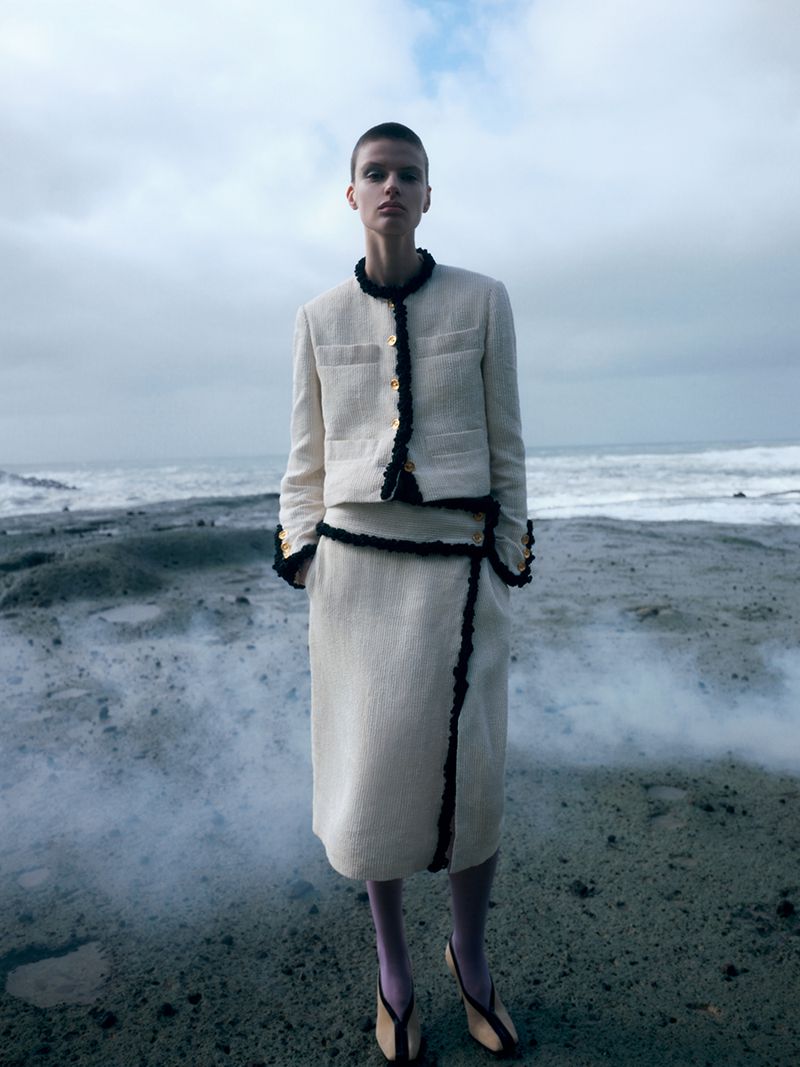 Model with short hair wearing a cream-colored textured skirt suit with black trim on a rocky shore by the ocean.