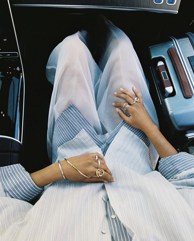 High-angle shot of a person seated in a vehicle wearing a striped shirt and sheer white skirt, adorned with rings and bracelets.