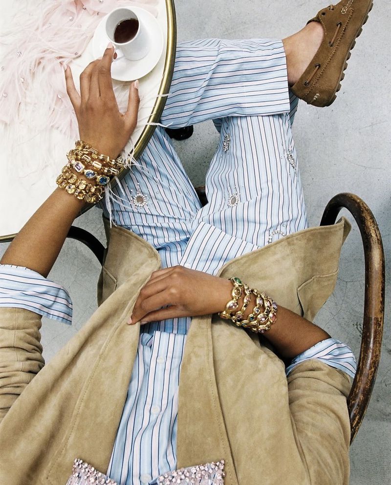 Overhead shot of a person wearing stacked gold bracelets, striped trousers, and brown suede loafers, holding a small coffee cup.