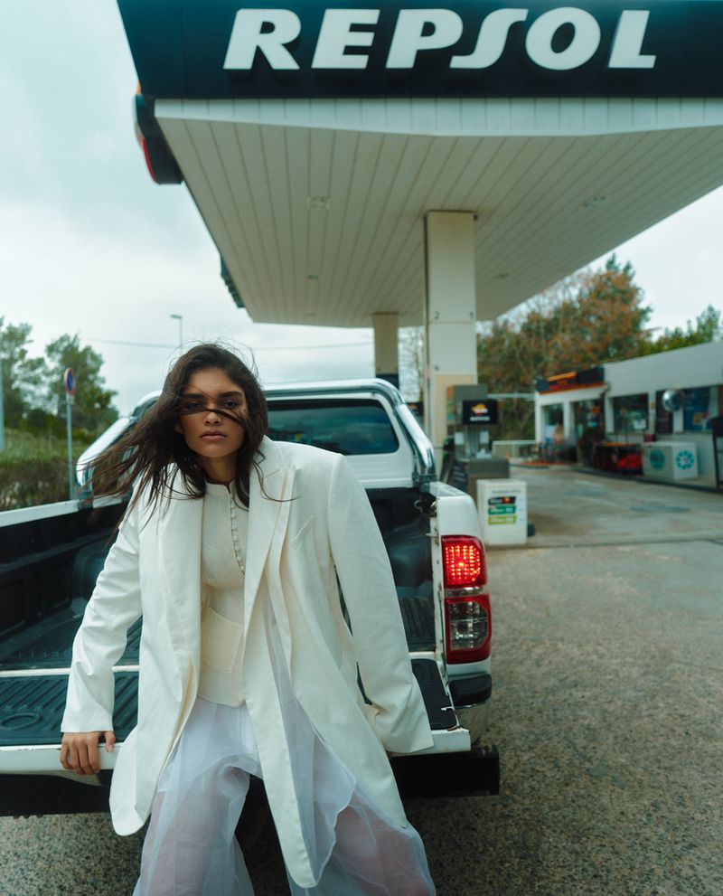Model in white blazer leaning against a white pickup truck at a Repsol service station.