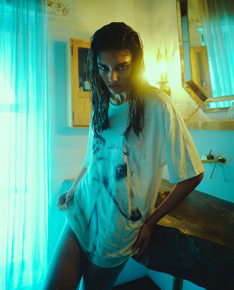 Model with wet dark hair wearing an oversized graphic t-shirt leaning against a basin in a turquoise lit room.